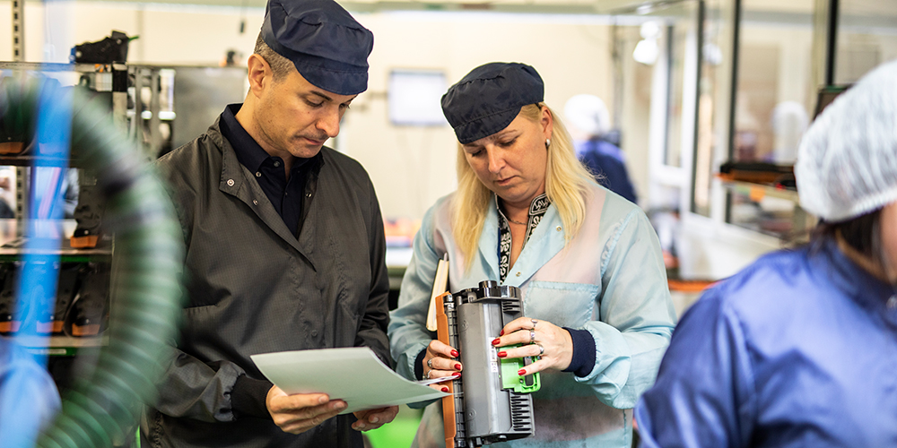 <p>Man and woman in factory setting surrounded by machinery and looking at a Brother toner cartridge.</p>