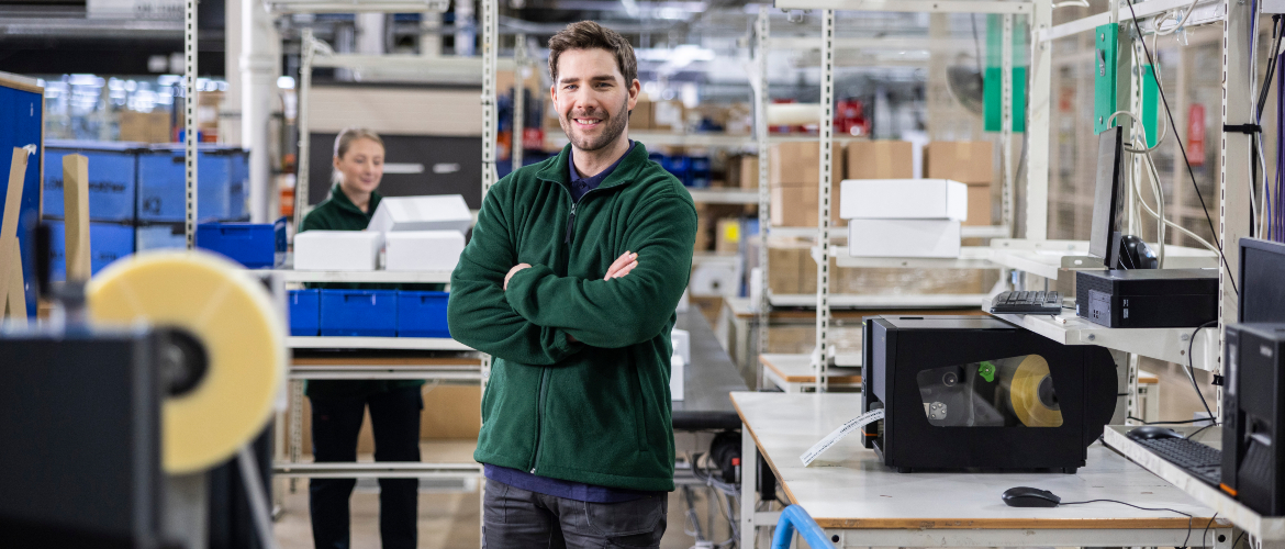 <p>Man and woman stood in a warehouse next to a labelling machine with boxes in the background</p>
