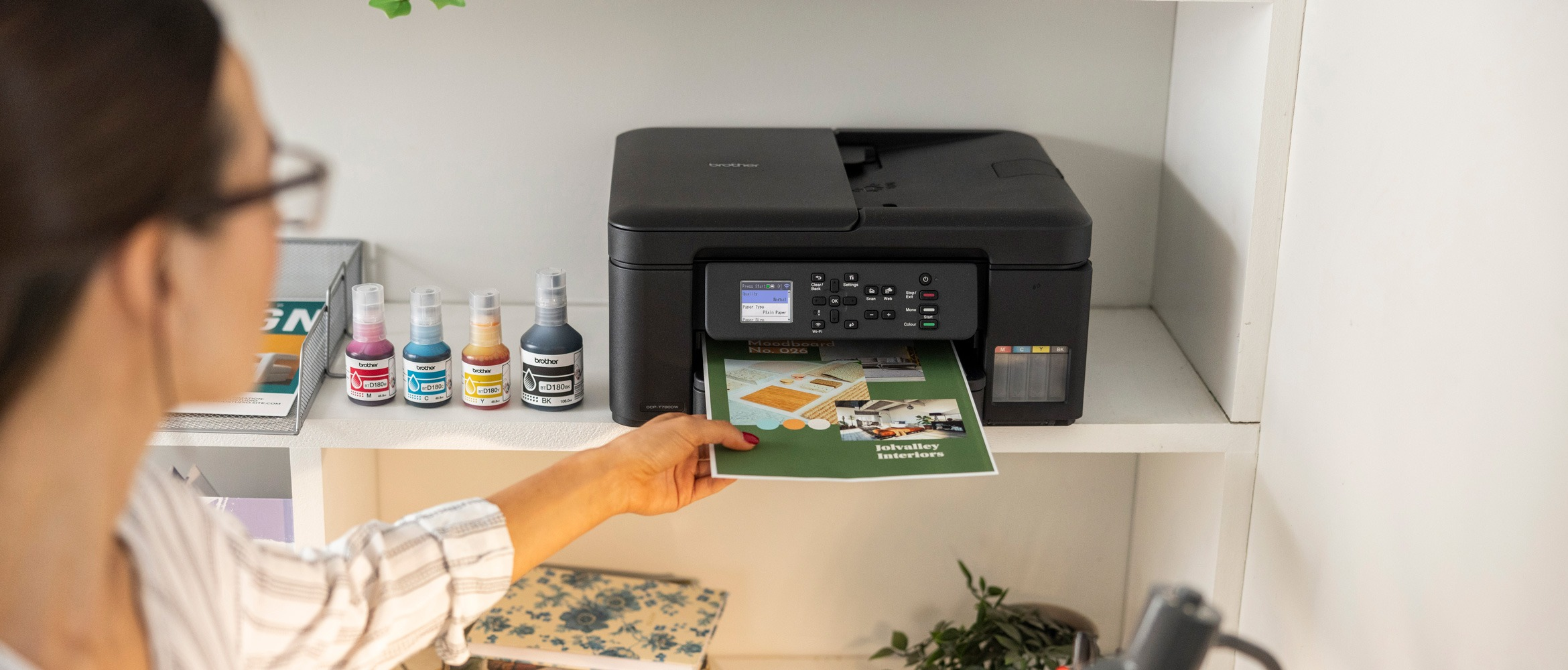 <p>A person standing in a bright home office, holding a printed poster next to a Brother inkjet printer, with plants on the windowsill and a long desk filled with printed materials, stationery, and laptop.</p>