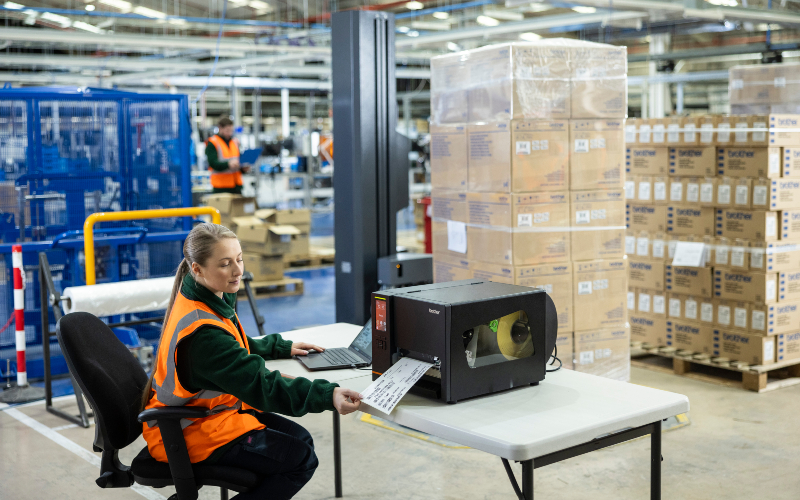 <p>Woman sat down at a table with a laptop, printing a label with stacks of boxes on pallets in the background</p>