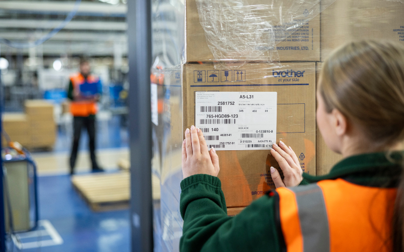 <p>Woman in a high-vis vest in a warehouse applying a label to a box</p>