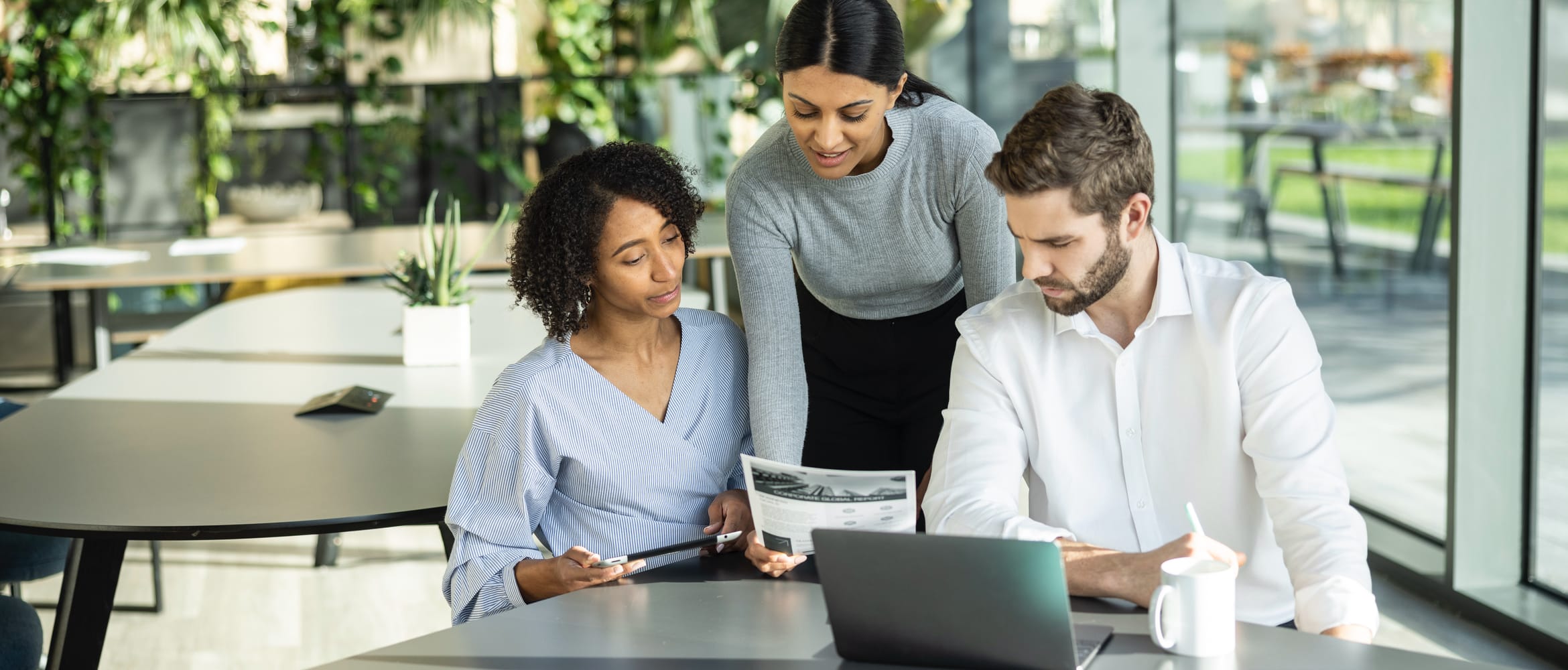 <p>Three business people reviewing a printed document while gathered around a desk in an open plan office</p>