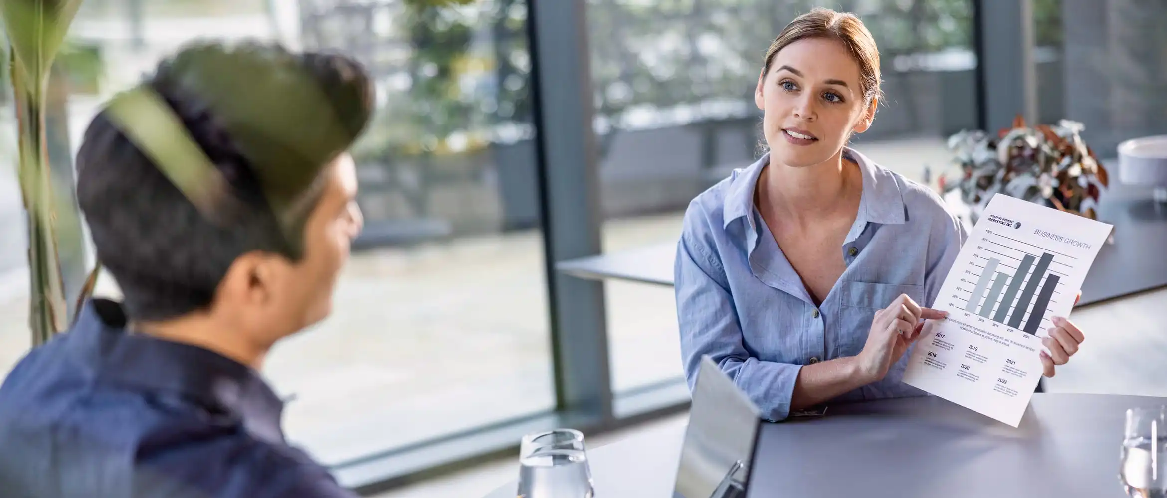 <p>Two business people looking at a printed chart while sat at a desk in an office environment</p>