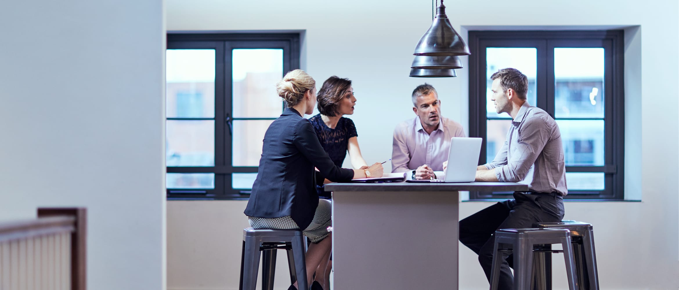 <p>SMB employees in a meeting, sat on stools around a high table</p>