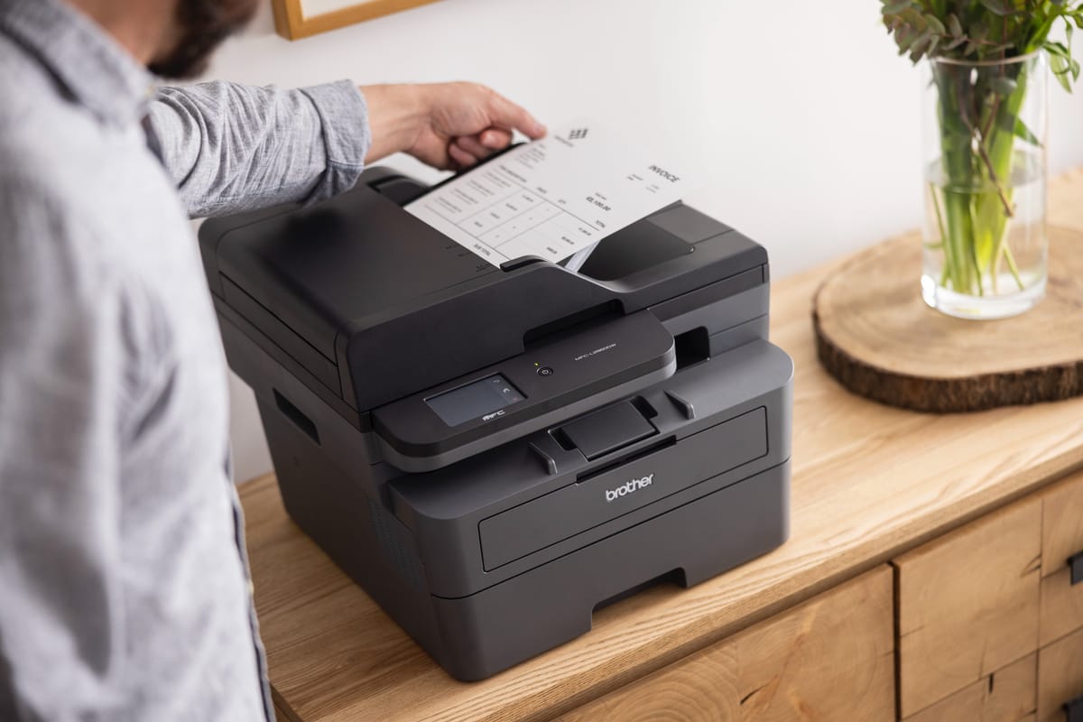 <p>A man retrieving print output from a Brother MFC-L2860DW mono laser printer which is on a wooden sideboard in a home environment</p>