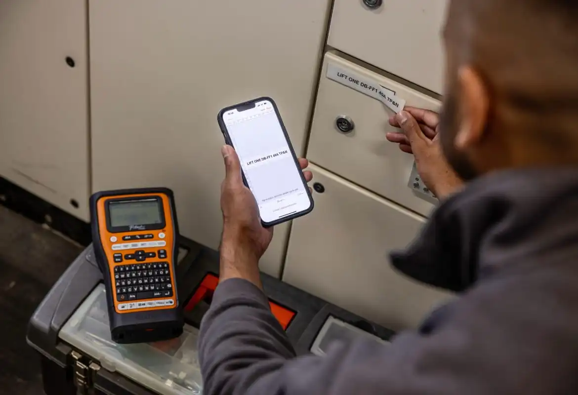 An electrician using Brother's Pro Label Tool app on a smartphone to print to a P-touch E-series label printer