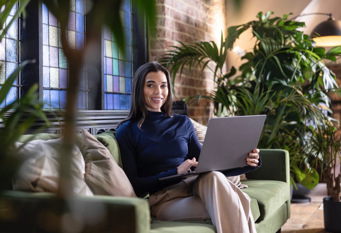 <p>A woman using a notebook computer white sat on a green velvet sofa with plants on either side and a stain glass window and exposed brickwork behind her</p>