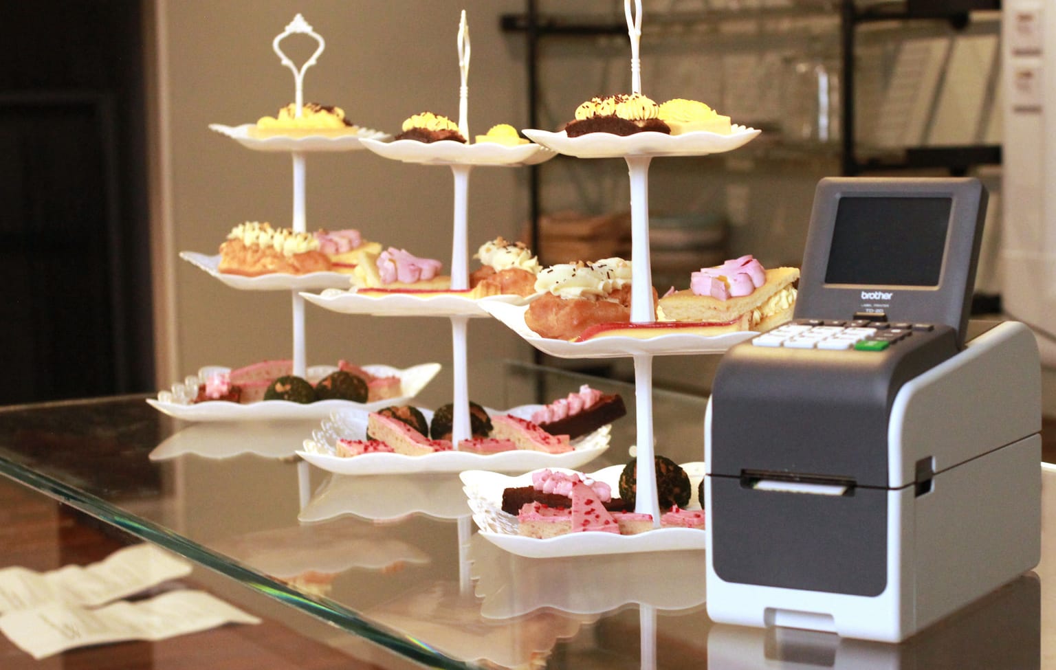 <p>Three cake stands containing an assortment if pastries on a glass surface next to a Brother TD-2D label printer</p>